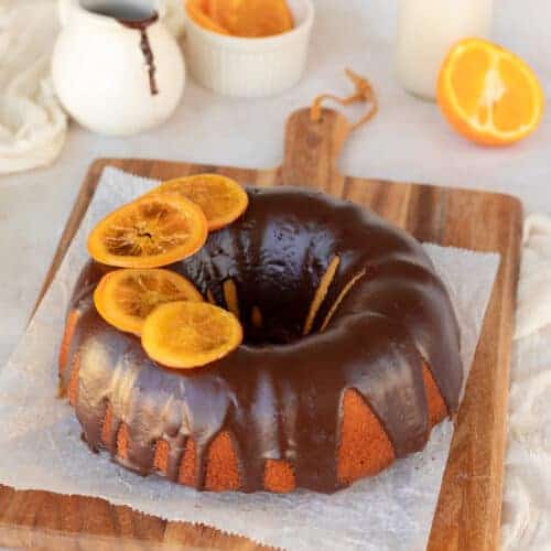 Orange bundt cake served on a wooden board and topped with chocolate ganache and candied orange slices.