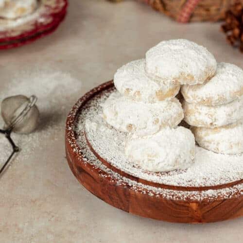 Kourampiedes stacked on a wooden stand topped with powdered sugar. Next to the stand there is a powdered sugar sifter.