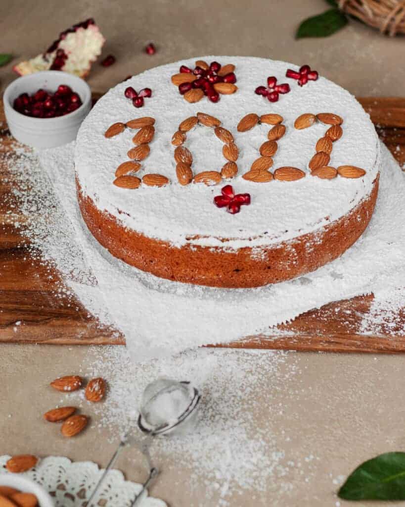 Shot of Greek vasilopita cake on a wooden board surrounded by pomegranate seeds and almonds.