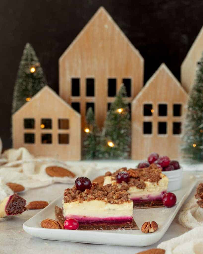 A slice of cranberry cheesecake bars on a platter. In the background a christmas house decoration with lights.