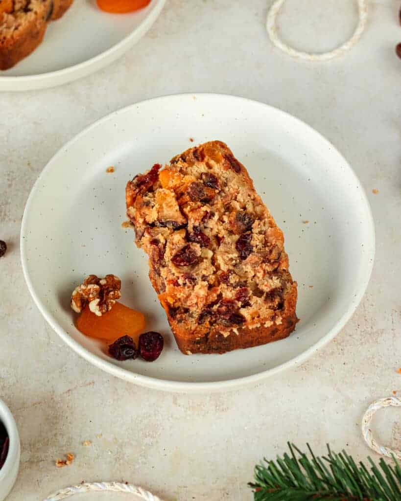 A slice of rum fruit cake served on a dessert plate with raisins, apricot and walnut.